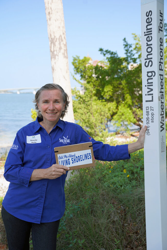 mandy baily standing next to a living shoreline in Sarasota with a "Ask Me About Living Shorelines" sign