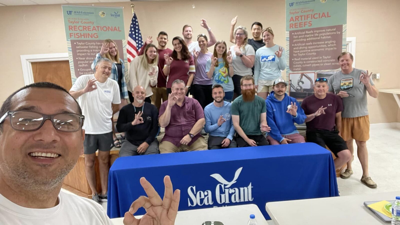 A group of about 15 people pose together indoors behind a blue Sea Grant table, with many making an OK hand sign.