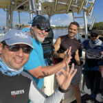 four scuba divers smile and pose together on a boat docked at a marina, with several making the OK hand signal.