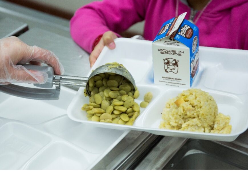 a cafeteria tray with a carton of milk, rice and a gloved hand emptying a scoop of lima beans onto the tray