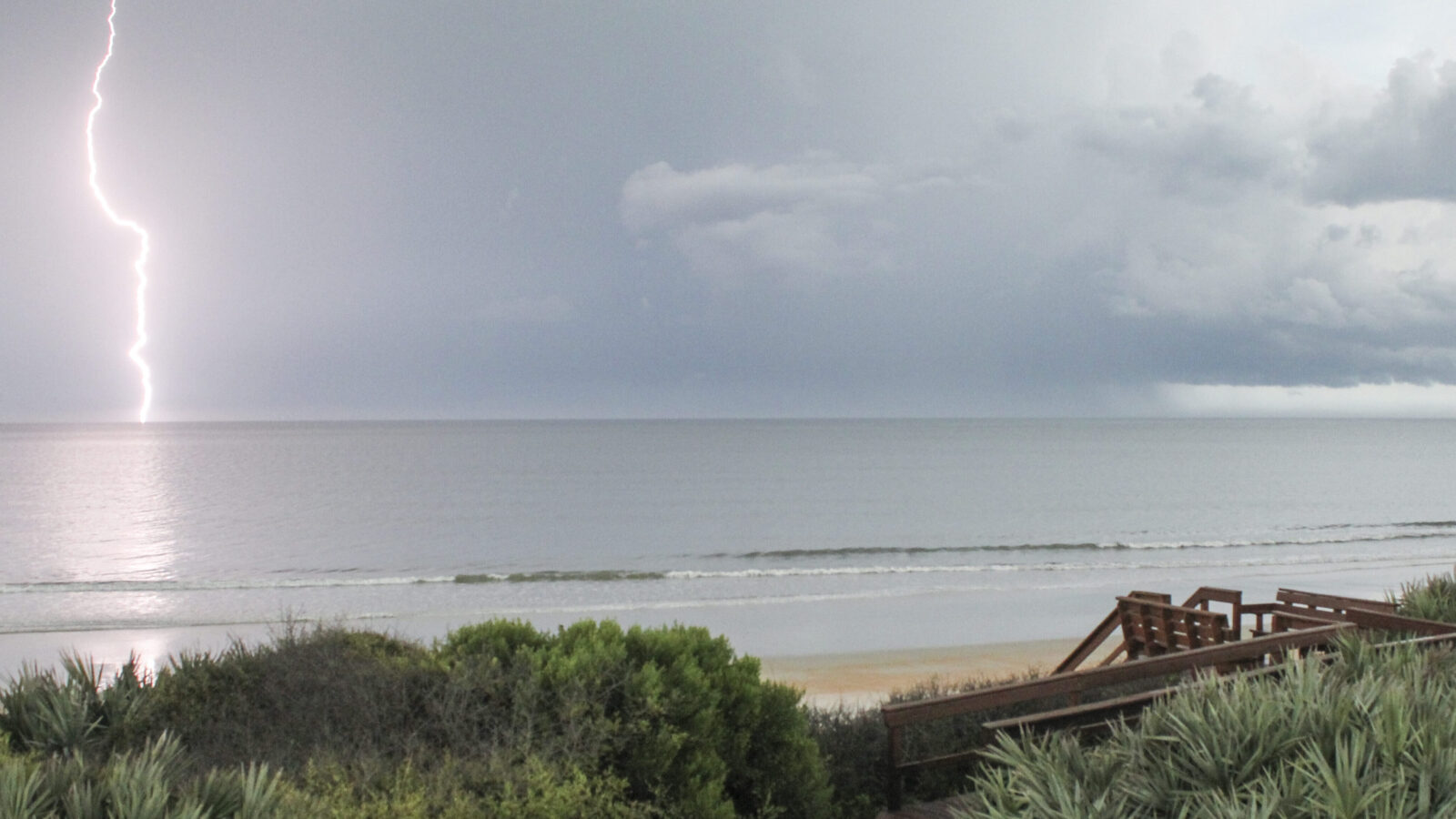 A single, bright bolt of lightning strikes the calm ocean horizon under a dark, stormy sky filled with heavy grey clouds. In the foreground, green coastal bushes and a wooden boardwalk structure overlook the sandy beach and gentle waves.