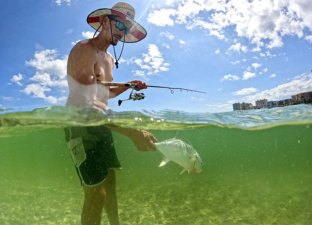 A split-level over-under shot shows Mike Sipos, UF/IFAS Extension Agent for Collier County standing in shallow, green-tinted water while holding a fishing rod. Above the water, he's wearing sunglasses and a wide-brimmed straw hat against a sunny, blue sky with white clouds. Below the surface, he holds a silvery fish by its tail over a sandy seabed.