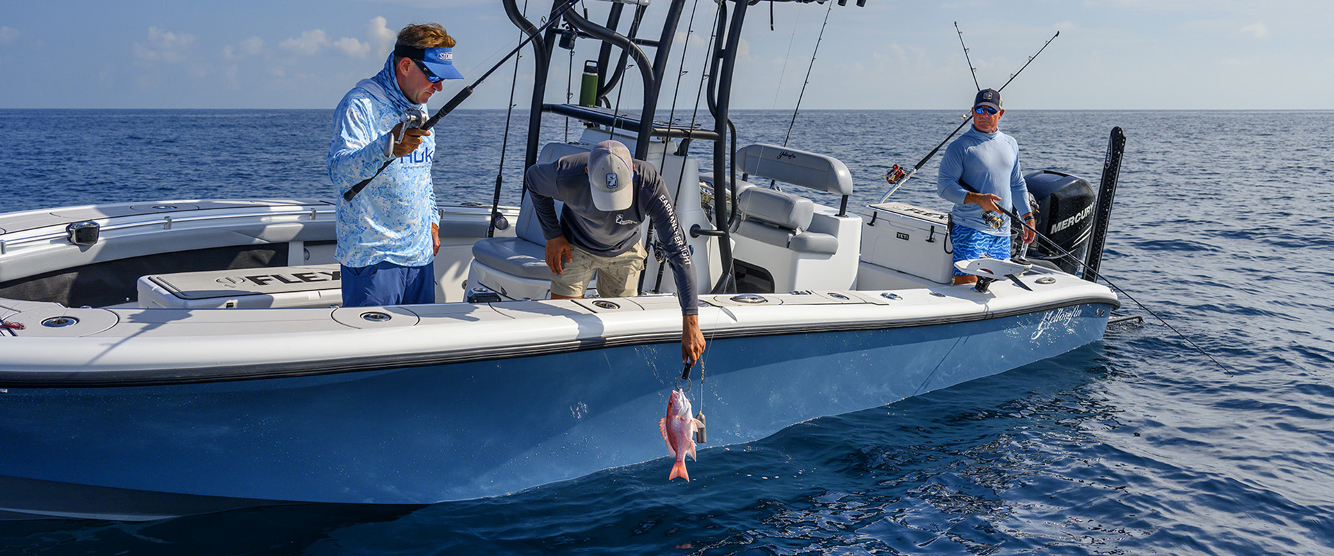 Three men fish from a light blue center-console boat on the open ocean under a clear sky. One man leans over the side of the boat, using a weighted line to lower a bright pink fish back into the water, while his two companions watch with their fishing rods in hand