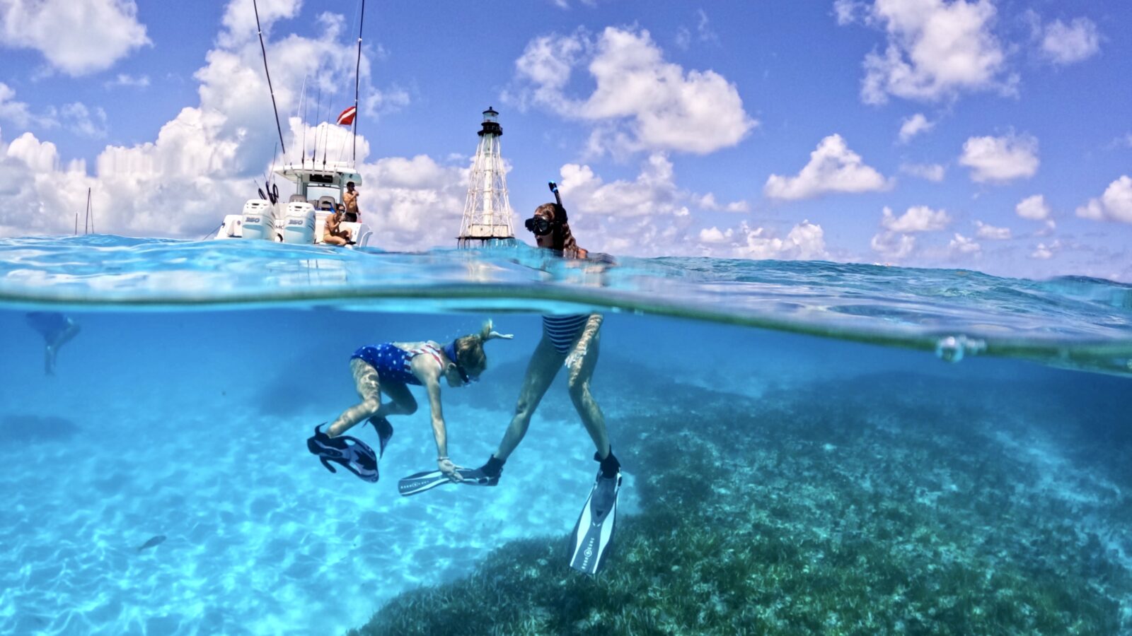 A split-level over-under shot shows two snorkelers swimming in clear turquoise ocean water over a shallow coral reef. Above the water line, a white boat sits anchored near a distant skeletal-frame lighthouse under a bright blue sky filled with puffy white clouds