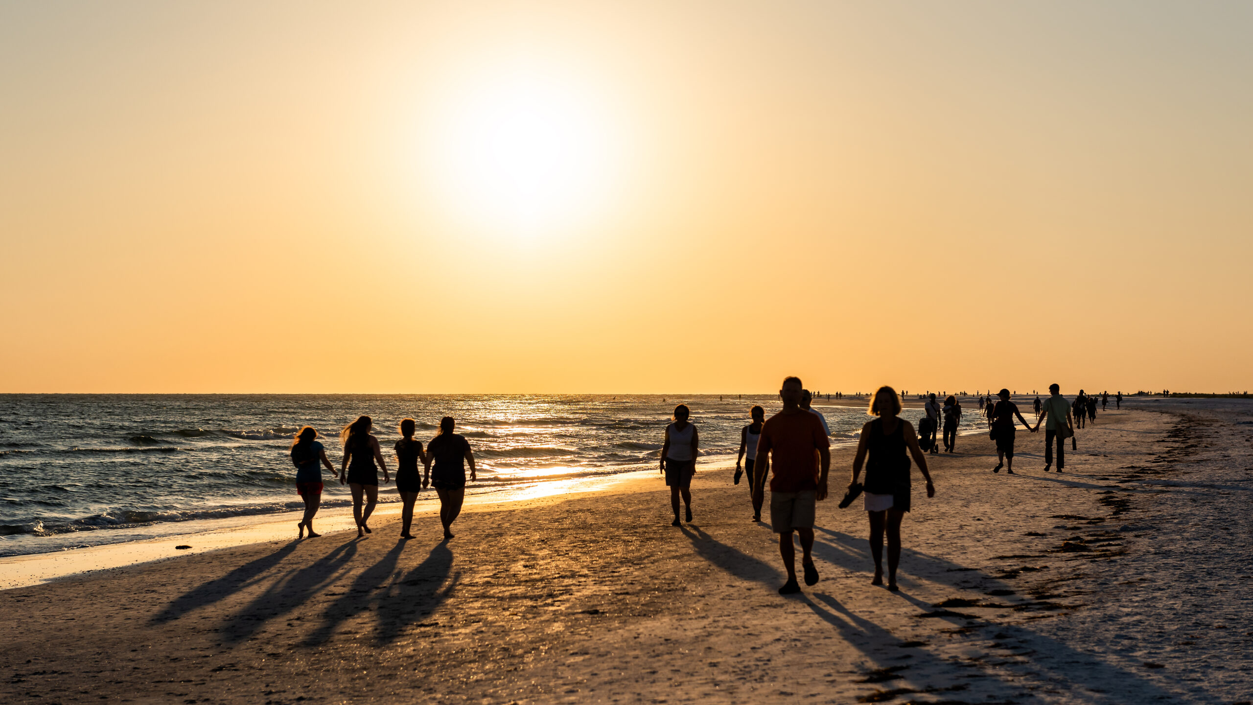 Silhouetted crowds of people stroll along a wide sandy beach during a bright sunset. The large, brilliant sun hangs low in a hazy golden sky, casting a long shimmering reflection across the ocean surface and stretching dark shadows from the walkers across the shoreline.