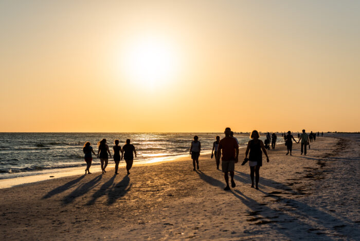 Silhouetted crowds of people stroll along a wide sandy beach during a bright sunset. The large, brilliant sun hangs low in a hazy golden sky, casting a long shimmering reflection across the ocean surface and stretching dark shadows from the walkers across the shoreline.