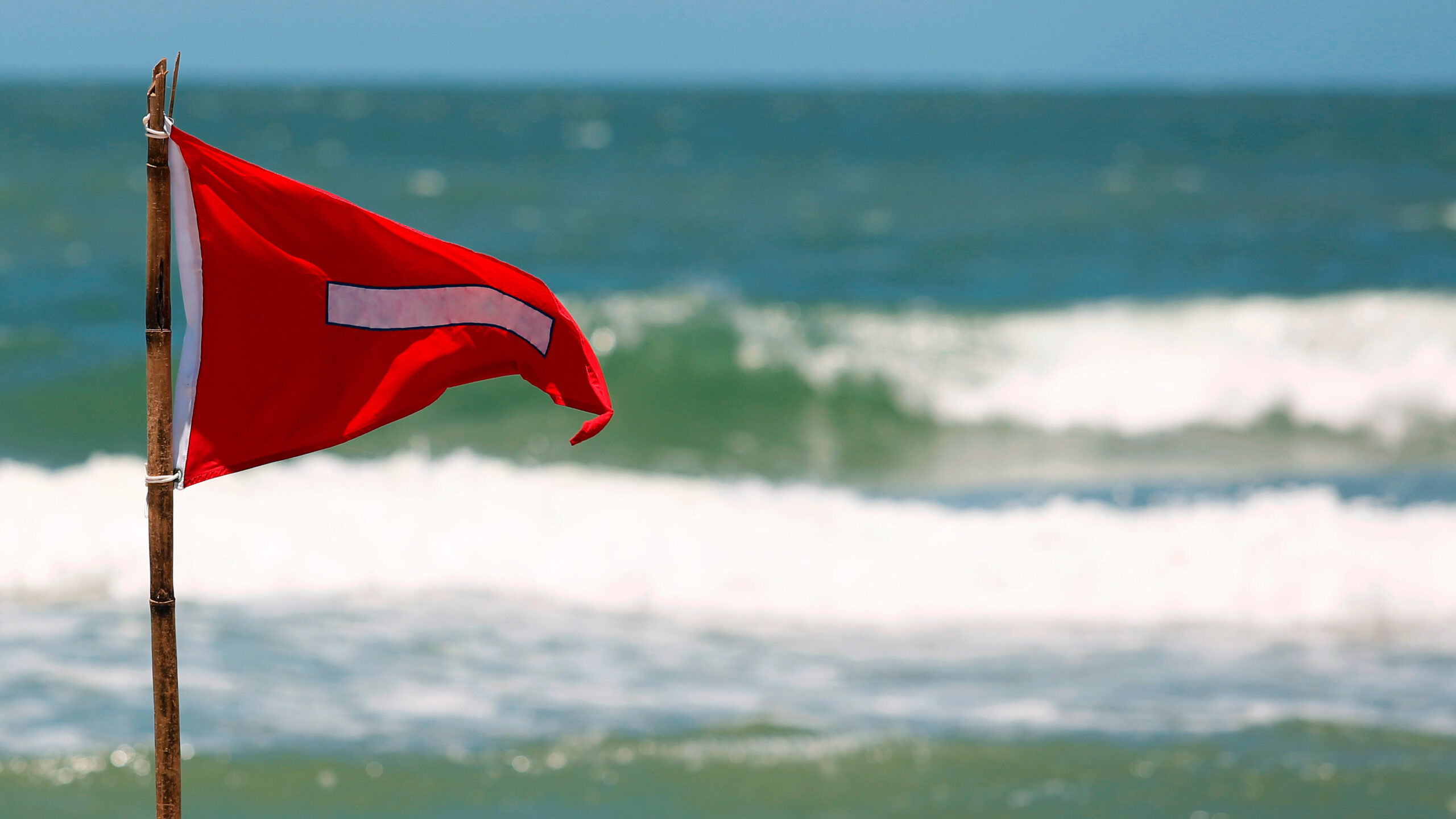 A bright red beach warning flag with a white horizontal stripe flies from a rustic wooden pole planted in the sand. The flag is positioned on the left side of the frame, set against a blurred background of teal ocean water, rolling white breaking waves, and a pale blue sky