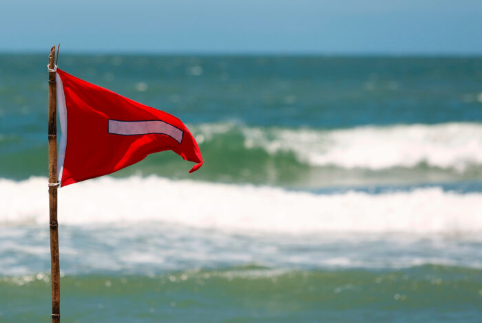 A bright red beach warning flag with a white horizontal stripe flies from a rustic wooden pole planted in the sand. The flag is positioned on the left side of the frame, set against a blurred background of teal ocean water, rolling white breaking waves, and a pale blue sky