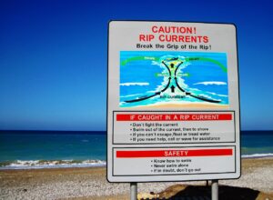 A large white safety sign with a black border stands on a sandy beach, warning visitors about rip currents under a clear blue sky. The sign features red and black text, an illustrated diagram of ocean currents pulling away from the shore, and bulleted safety instructions, set against a background of the blue ocean and breaking white waves