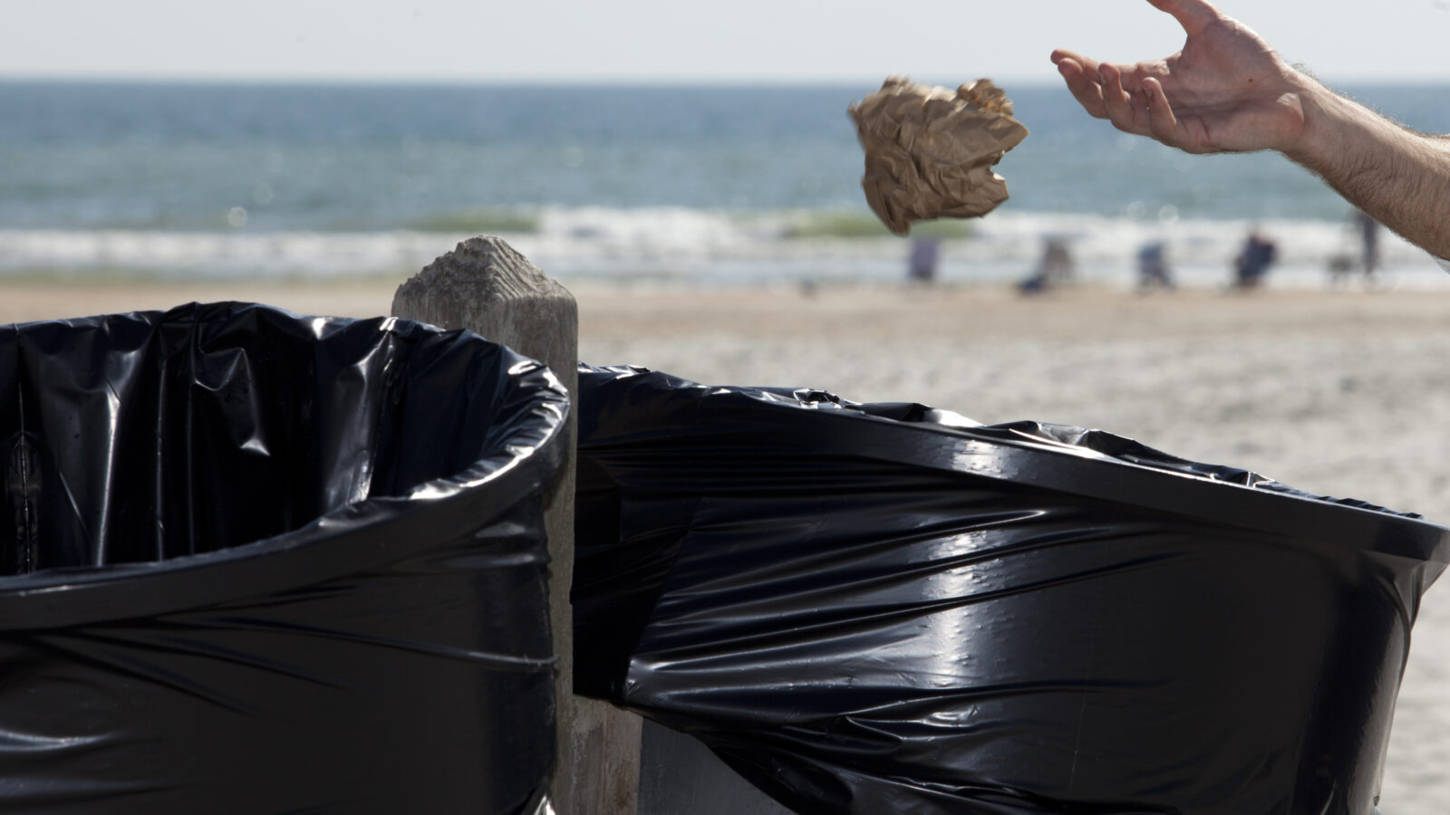 An arm extends from the right side of the frame, throwing a crumpled brown paper bag into one of two public trash cans lined with black plastic bags. The scene is set on a sunny beach, with a wooden post holding the bins and the ocean waves softly blurring in the background