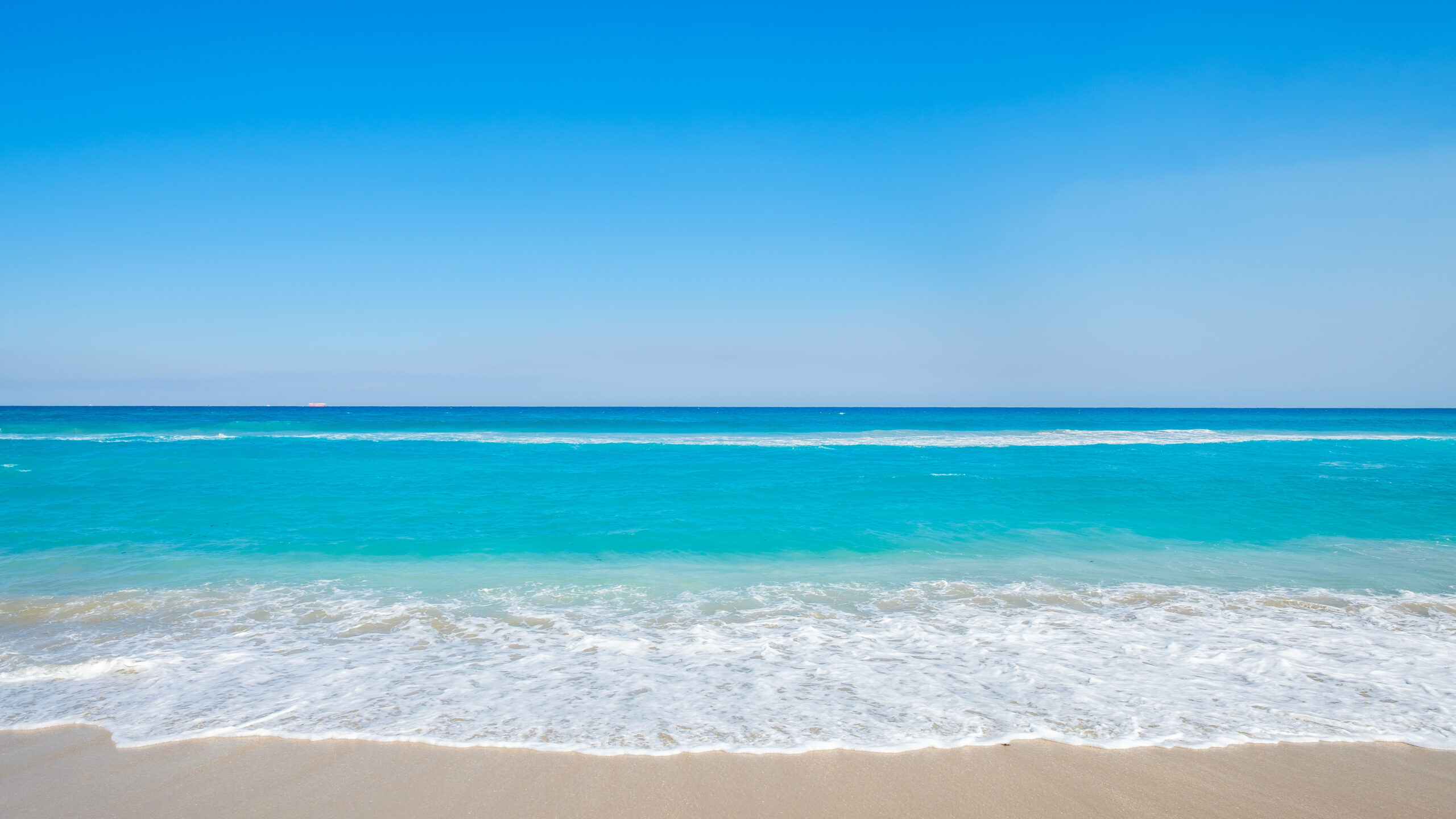 A wide, sunny shot captures a tranquil sandy beach meeting vibrant turquoise ocean waters under a vast, cloudless blue sky. White sea foam rolls gently onto the beige shoreline, creating a peaceful, minimalist coastal landscape.