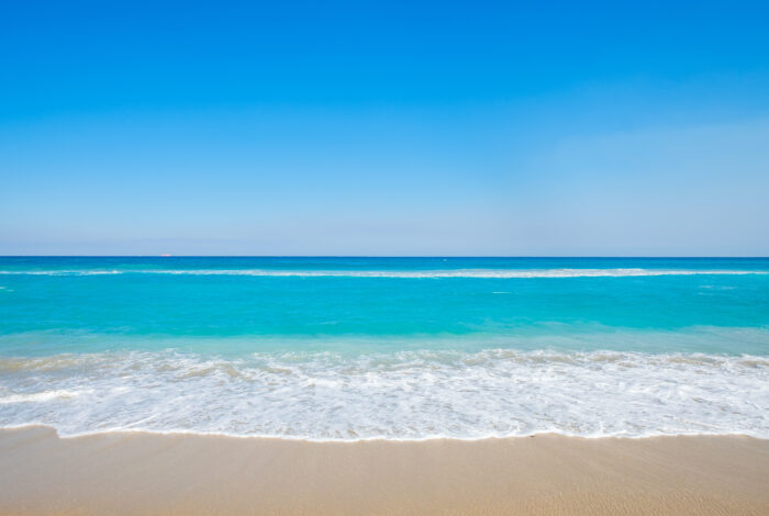 A wide, sunny shot captures a tranquil sandy beach meeting vibrant turquoise ocean waters under a vast, cloudless blue sky. White sea foam rolls gently onto the beige shoreline, creating a peaceful, minimalist coastal landscape.