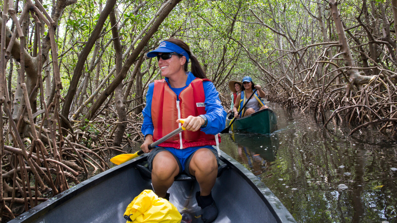 Pinellas County Extension agent Libby Carnahan wearing a blue shirt and red life jacket paddles a canoe down a narrow waterway lined with dense mangrove roots, followed by another canoe with two people at Weedon Island Preserve in Clearwater, Florida