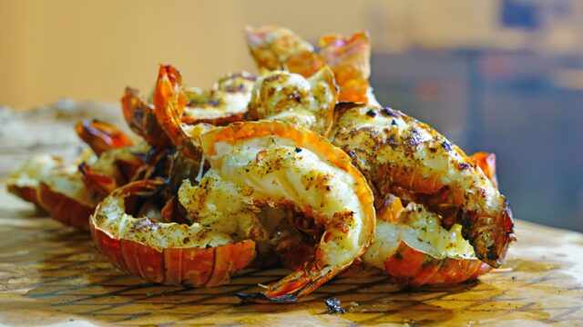 A pile of grilled spiny lobster tails rests on brown butcher paper. The meat is split open, showing a white interior seasoned with black pepper and herbs, while the outer shells are a vibrant orange-red with dark char marks from cooking