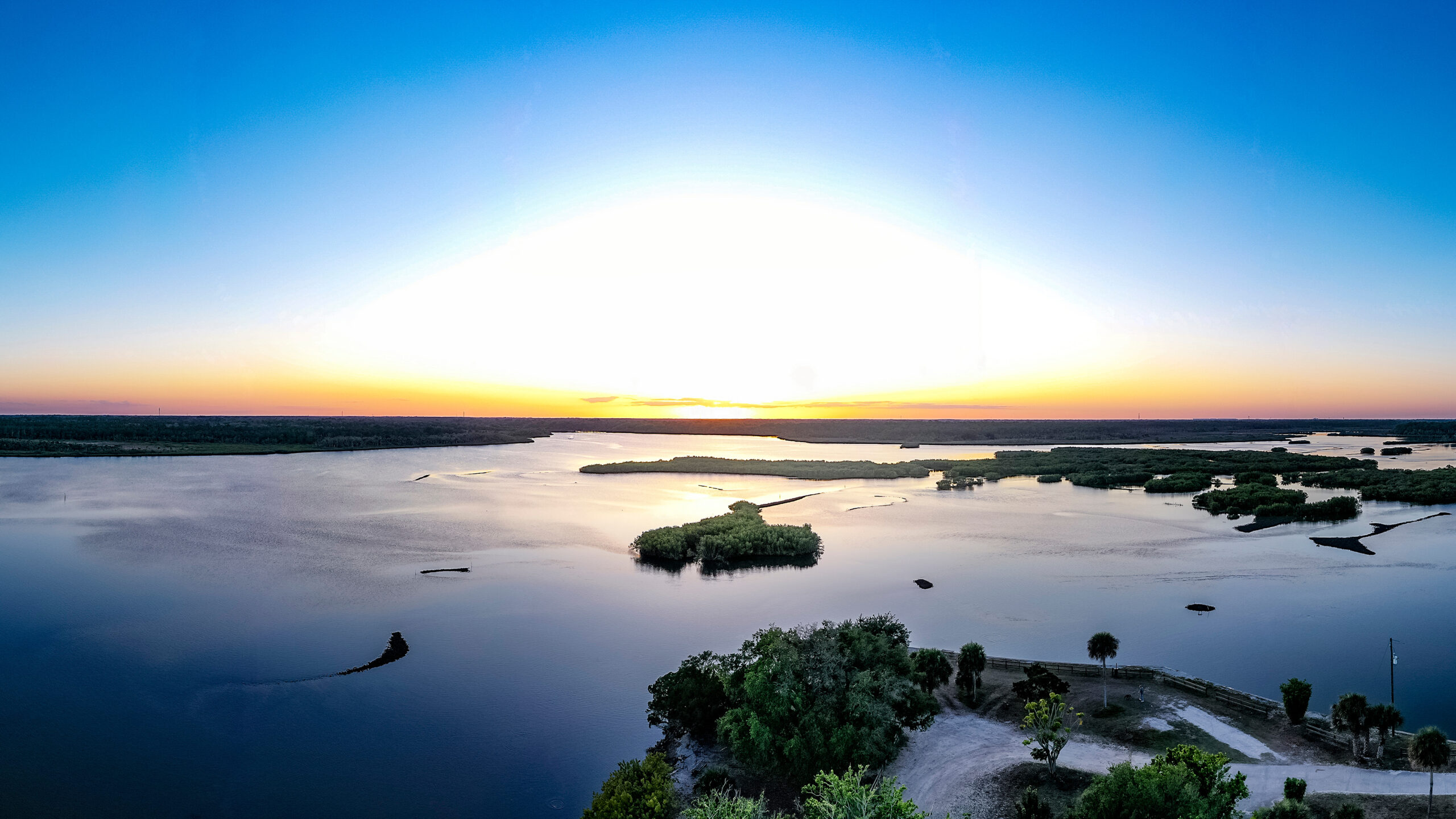 A wide panoramic aerial view shows a bright sunset casting a golden glow over Spruce Creek watershed, New Symrna Beach, Florida, USA, dotted with small islands and dense shoreline vegetation.