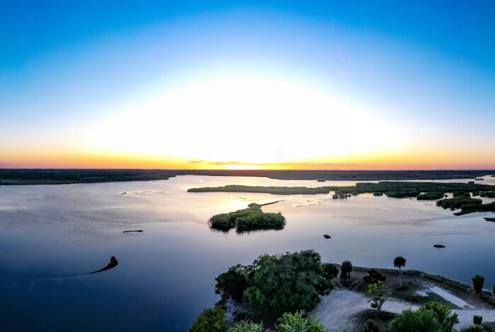 A wide panoramic aerial view shows a bright sunset casting a golden glow over Spruce Creek watershed, New Symrna Beach, Florida, USA, dotted with small islands and dense shoreline vegetation.