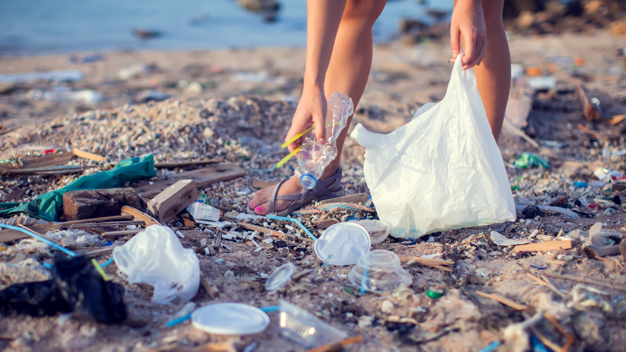 A person bends down on a heavily littered sandy beach to pick up a crumpled plastic bottle, holding an open white plastic trash bag to collect the surrounding debris.