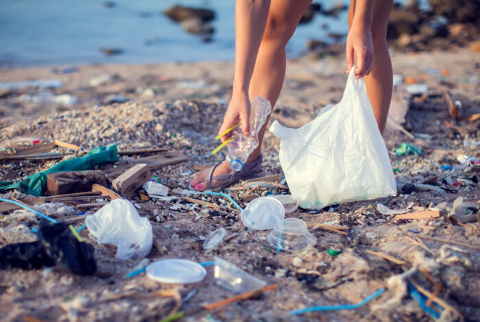 A person bends down on a heavily littered sandy beach to pick up a crumpled plastic bottle, holding an open white plastic trash bag to collect the surrounding debris.