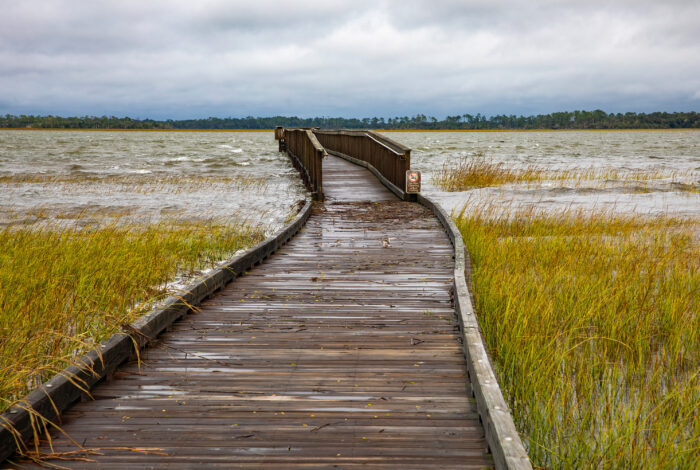 A flooded wooden boardwalk curves forward into a vast coastal marshland under a cloudy, overcast sky.
