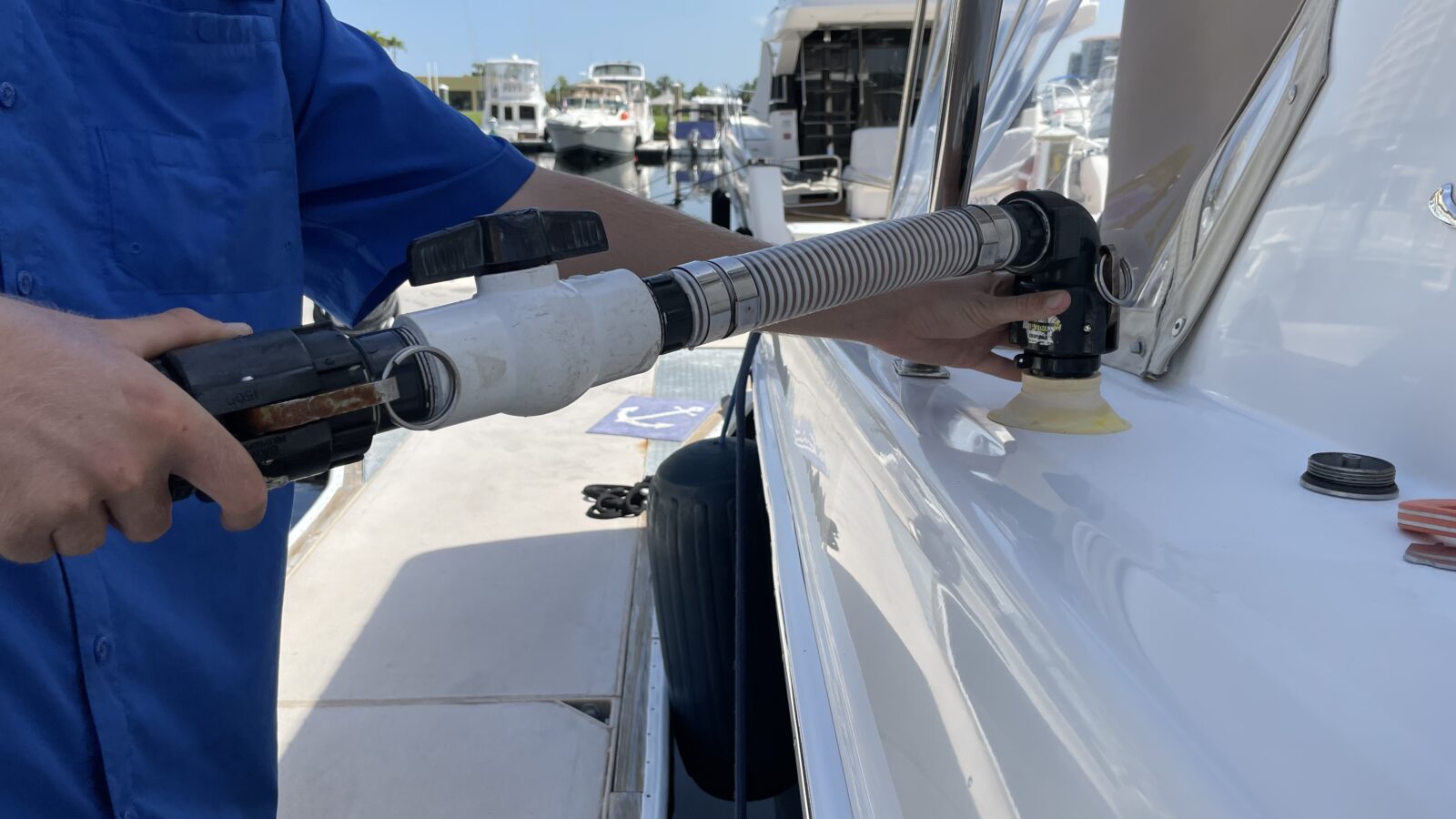 A close-up shot shows a person's hands connecting a flexible, heavy-duty waste pumpout hose to a discharge fitting on the side of a white boat docked at a marina.