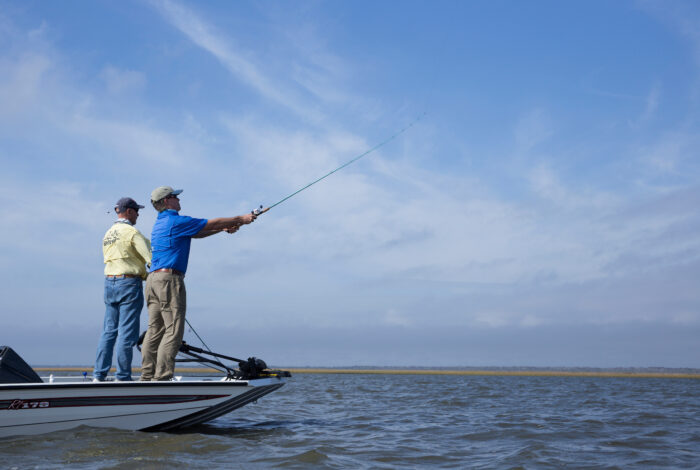 Two anglers standing on the bow of a motorboat casting a fishing rod into the water under a clear blue sky