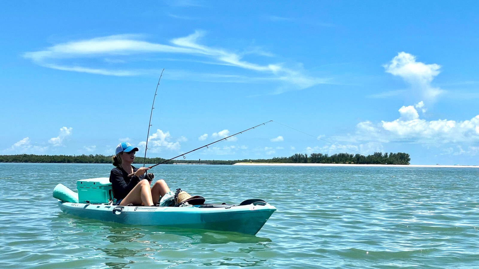A person wearing a blue cap and sunglasses sits in a light-blue fishing kayak on calm, turquoise water under a bright blue sky with wispy clouds. The kayaker holds a fishing rod extending over the water, with a teal cooler strapped to the back of the vessel. In the distant background, a small, sandy island with green trees sits along the horizon line.