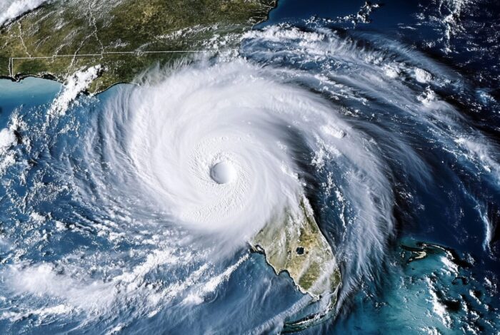 An aerial satellite view shows a massive, swirling hurricane with a well-defined circular eye positioned over the Gulf of Mexico and the Florida peninsula. The bright white, counter-clockwise spiral of thick clouds covers most of Florida and extends across the deep blue ocean water toward the southeastern United States coastline