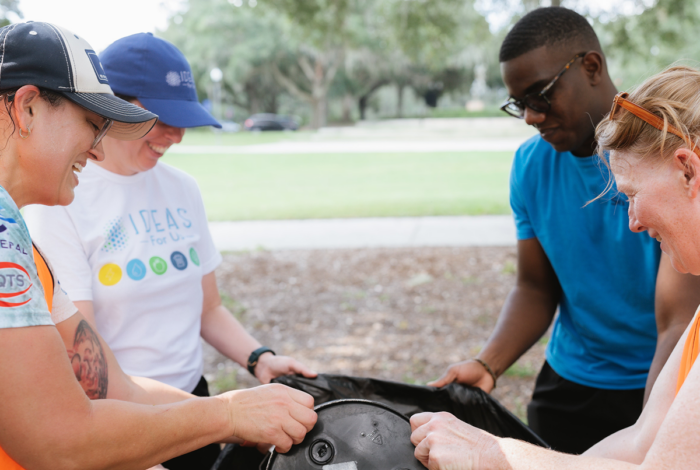 outdoor photo of two people holding a trash bag open and two more people dumping a plastic bucket into the bag