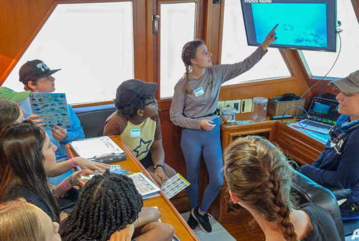 Sophia points at a screen while demonstrating her research as a group of young onlookers observes.