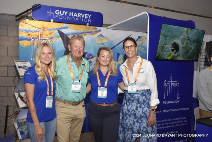 Two 2024 FSG-Guy Harvey Fellow posing with Dr. Guy Harvey and his daughter, Jessica Harvey, in front of banner.