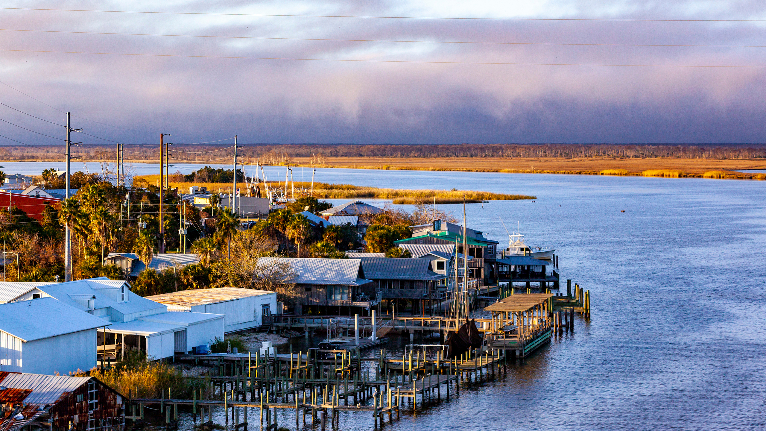 An elevated, wide-angle view of a coastal waterfront town under a dramatic, cloudy sky.On the right, a wide, calm river or bay flows horizontally across the frame. On the left bank, a cluster of rustic, metal-roofed houses, wooden docks, and piers extend into the water. A tall mast from a docked sailboat rises near the center. Palm trees and green foliage are scattered between the buildings and utility poles. In the background, across the water, a flat expanse of golden marshland stretches beneath a thick layer of low, heavy storm clouds rolling across the horizon