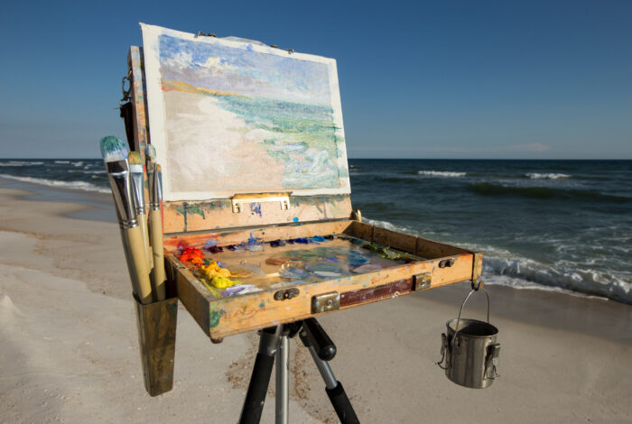 An artist's wooden plein air easel stands on a sandy beach, holding a partially completed oil painting that depicts the surrounding coastal landscape. The paint-splattered wooden box rests on a tripod stand, revealing an open palette covered in vibrant mounds of oil paint, with several long paintbrushes stored in a side container and a small metal bucket hanging from the opposite side. In the background, calm ocean waves roll onto the white sand shore under a clear blue sky, mirroring the scenic view captured on the canvas.